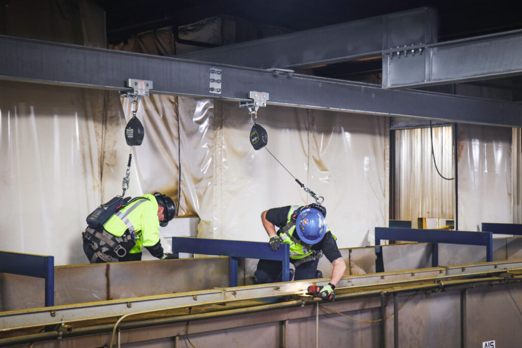 two workers on top of equipment attached with a harness to overhead fall protection beams inside warehouse
