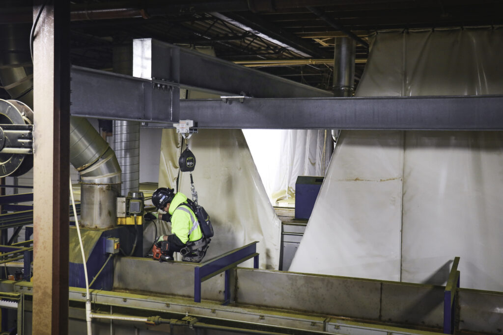 two workers on top of equipment attached with a harness to overhead fall protection beams inside warehouse