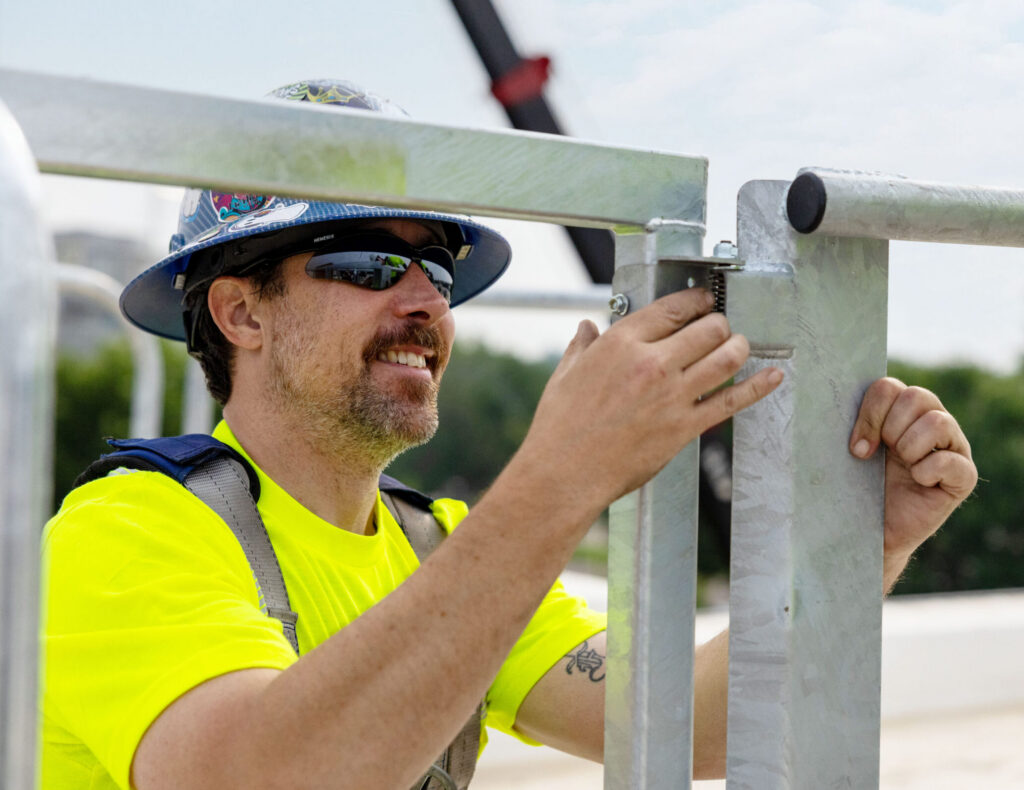Close-up view of an on-site installation of a spring-loaded, industrial-grade galvanized steel safety gate onto a modular edge protection system by a worker in full PPE.