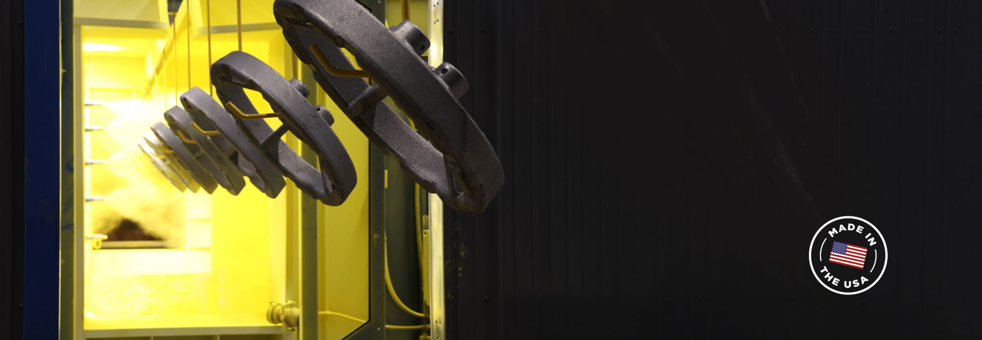 Dark metal industrial bases hanging from an overhead conveyor line in a brightly lit yellow processing booth, with a "Made in the USA" badge in the corner