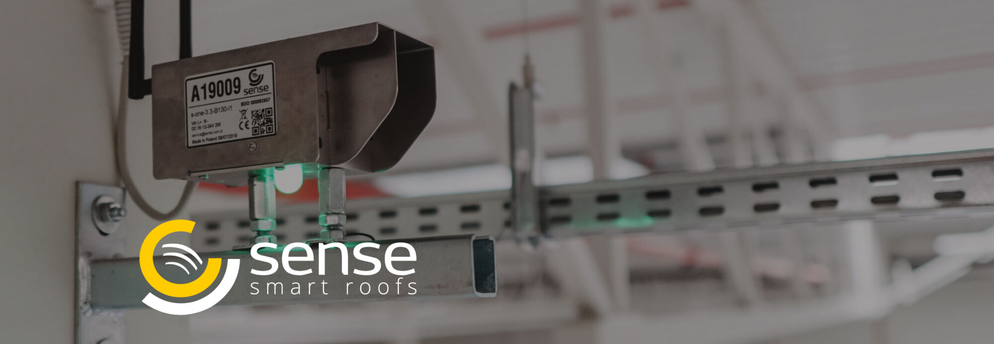 Close-up of a Sense Smart Roofs electronic sensor unit mounted on a ceiling support beam.Overlaid in the foreground is the Sense Smart Roofs logo featuring yellow and white concentric arches.