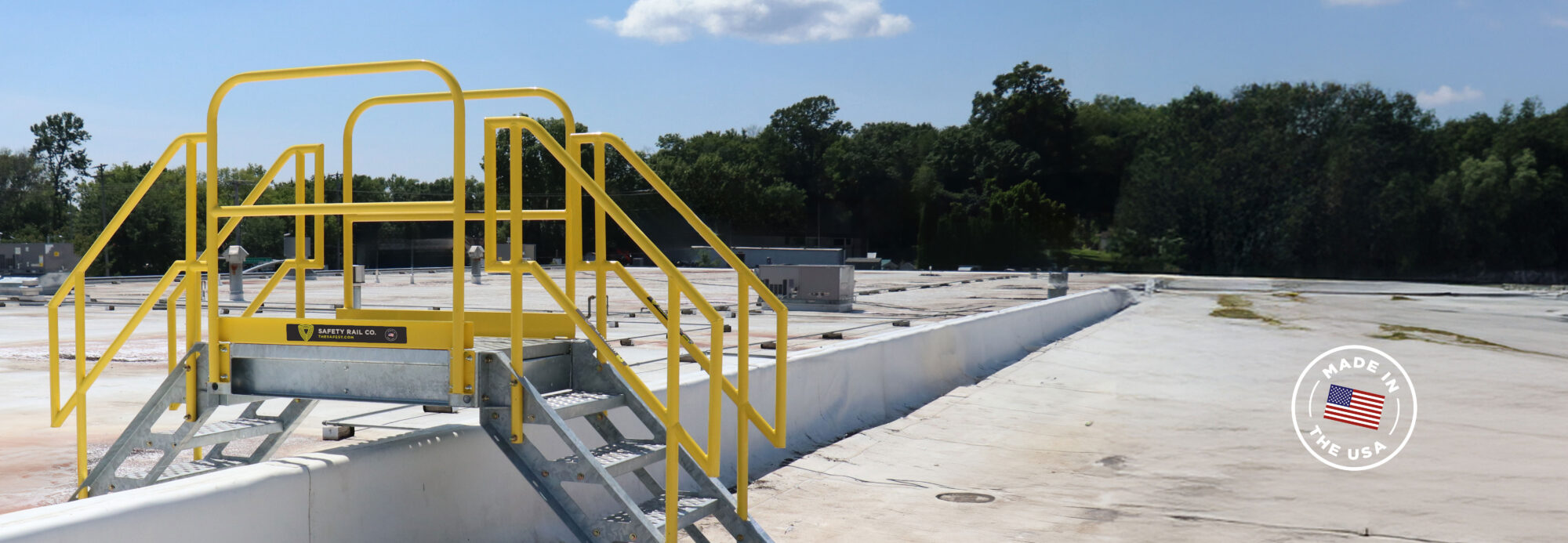 A metal crossover bridge featuring bright yellow safety railings and dual staircases by Safety Rail Co., bridging over a rooftop barrier on a expansive commercial building. A "Made in the USA" badge is visible in the bottom right corner.